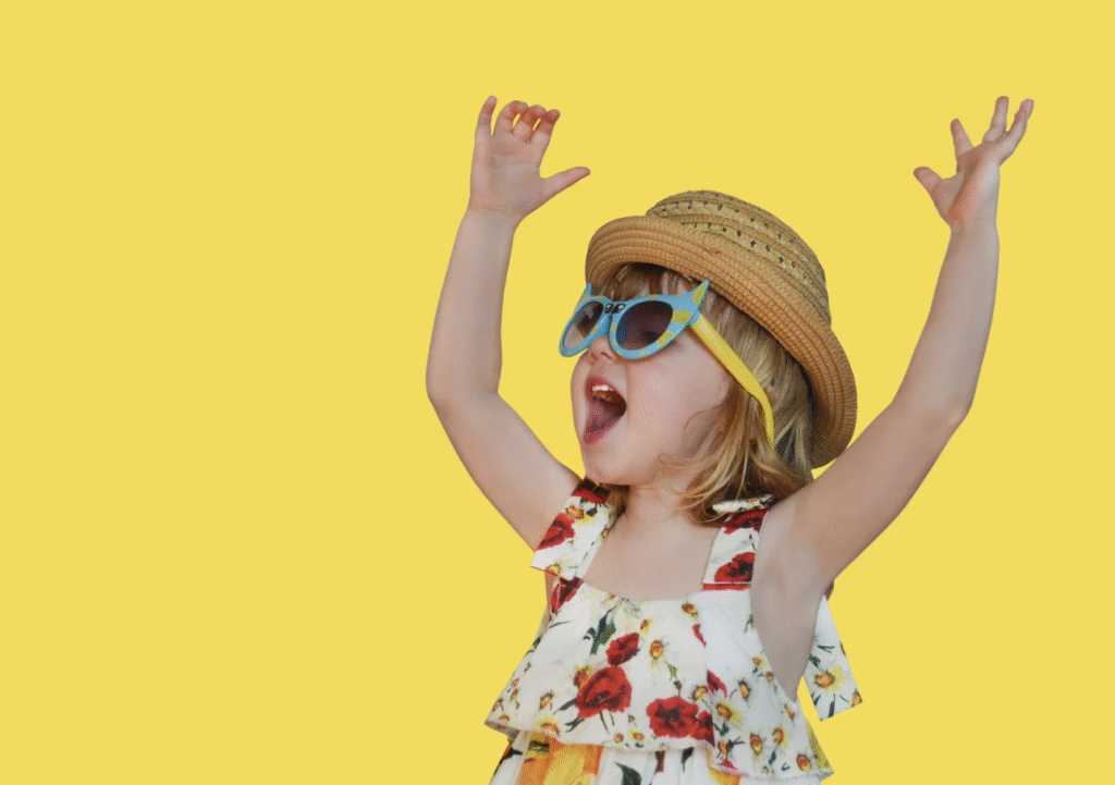 a young child wearing sunglasses and a hat has her arms raised in a joyful expression